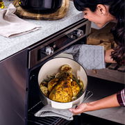 Frau nimmt einen ovalen Gusseisen Bräter von KitchenAid mit gebratenem Hähnchen und Gemüse aus dem Ofen. Seitenansicht.