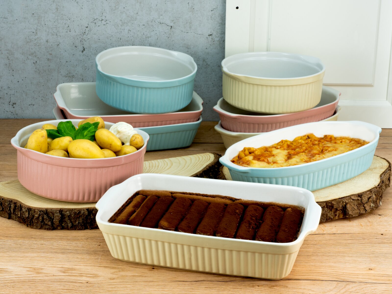 Pastellfarbene Keramik-Auflaufformen und runde Schalen mit Rillenstruktur, teils gestapelt vorne eine rechteckige Form mit Dessert, daneben eine Form mit Auflauf und eine Schale mit Kartoffeln – aufgenommen aus leicht erhöhter Frontperspektive.