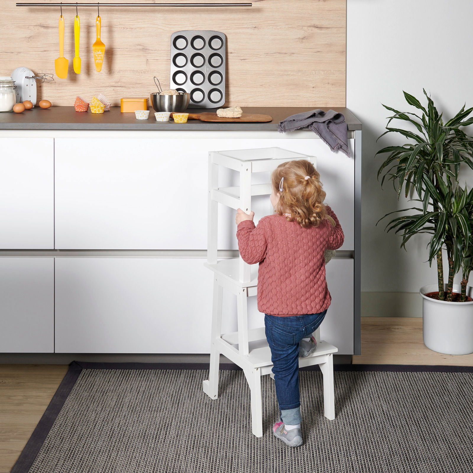 MyBaby Lernturm TOWER Ein Kind steht auf einem weißen Lernturm in einer Küche, um die Arbeitsfläche zu erreichen. Perspektive von hinten.