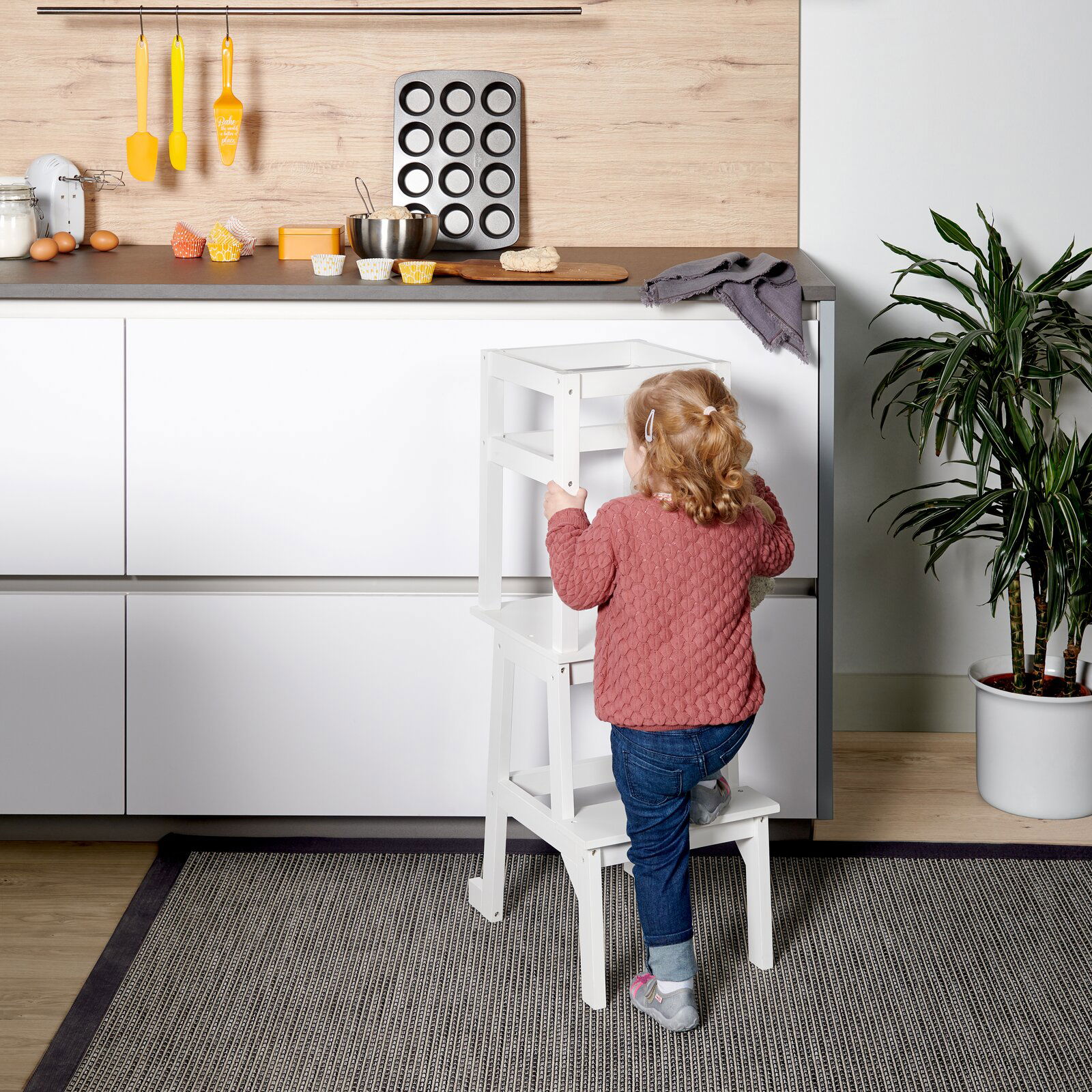 MyBaby Lernturm TOWER Ein Kind steht auf einem weißen Lernturm in einer Küche, um die Arbeitsfläche zu erreichen. Perspektive von hinten.