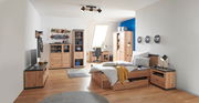 Gemütliches Schlafzimmer mit einem Holzbett, das von der Seite gezeigt wird. Das Bett ist mit gestreifter Bettwäsche und einem gelben Überwurf dekoriert. Im Hintergrund sind ein Schreibtisch, ein Kleiderschrank und Regale zu sehen.