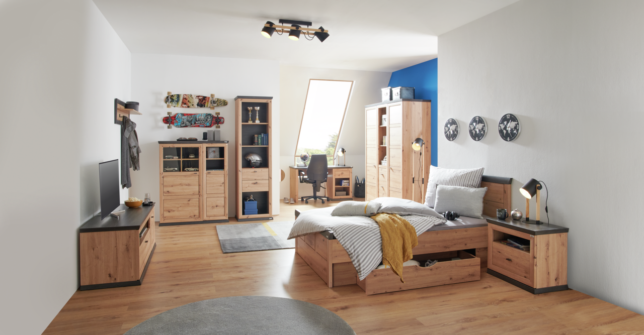 Gemütliches Schlafzimmer mit einem Holzbett, das von der Seite gezeigt wird. Das Bett ist mit gestreifter Bettwäsche und einem gelben Überwurf dekoriert. Im Hintergrund sind ein Schreibtisch, ein Kleiderschrank und Regale zu sehen.