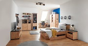 Een gezellige slaapkamer met een houten bed, dat vanaf de zijkant te zien is. Het bed is opgemaakt met gestreept beddengoed en een gele sprei. Op de achtergrond zijn een bureau, een kledingkast en planken te zien.