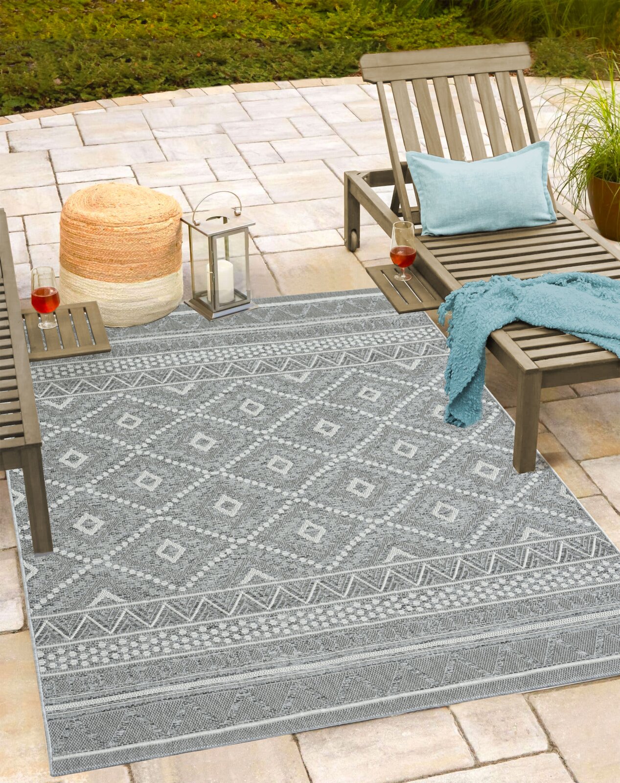 Grauer Outdoor-Teppich mit geometrischem Rauten- und Bordürenmuster auf einer Terrasse zwischen zwei Holzliegen, fotografiert aus leicht erhöhter schräger Perspektive.