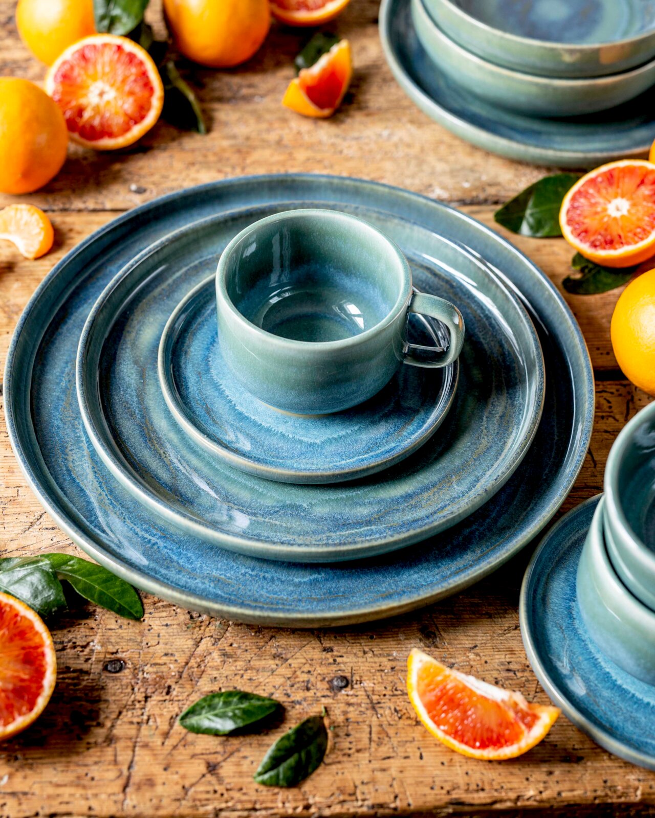 Gipfelstück Kaffeeservice 12-tlg. TIEFER SEE Blau-grünes Keramik-Geschirrset mit Tasse auf Teller und großem Speiseteller auf rustikalem Holztisch, umgeben von Blutorangen und grünen Blättern, aus leicht erhöhter schräger Draufsicht
