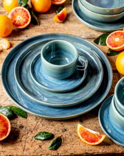 Blau-grünes Keramik-Geschirrset mit Tasse auf Teller und großem Speiseteller auf rustikalem Holztisch, umgeben von Blutorangen und grünen Blättern, aus leicht erhöhter schräger Draufsicht