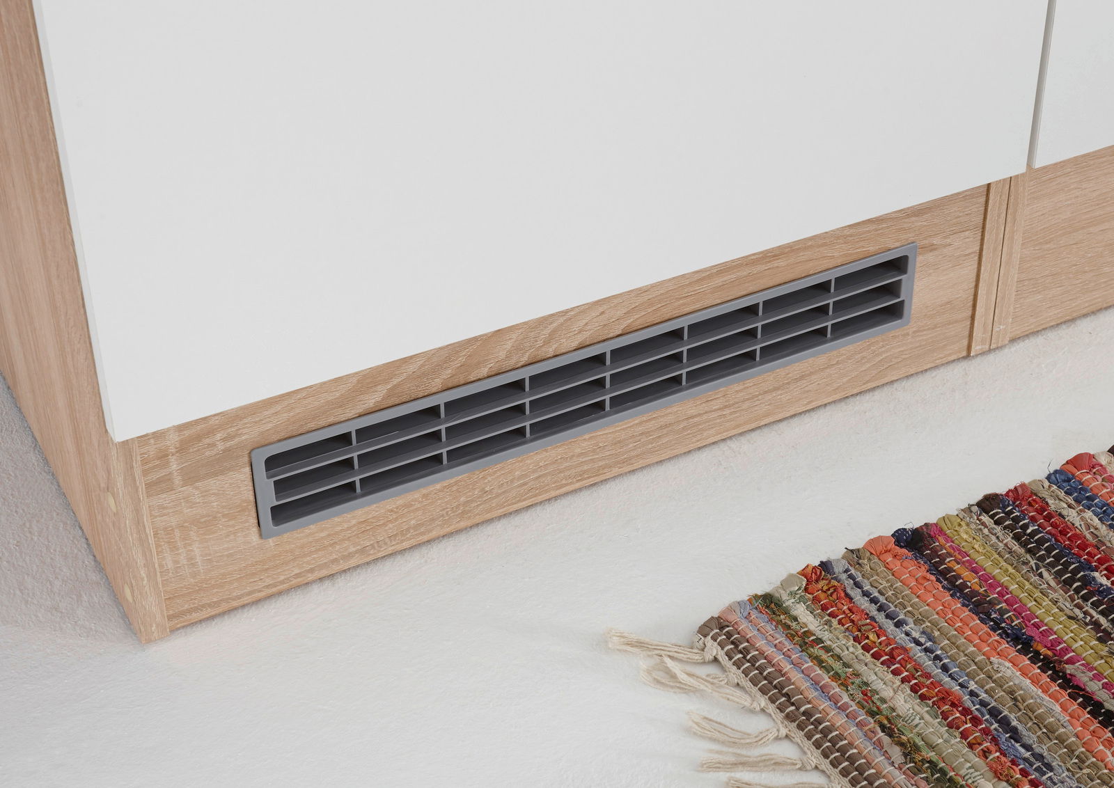 Nahaufnahme eines Küchenschranks mit Holzoptik und Lüftungsgitter, von vorne betrachtet, mit einem bunten Teppich am Boden.