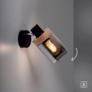 Moderne Wandlampe in Schwarz mit Holzdetail und zylindrischem Rauchglas-Schirm, leuchtendem Edison-Leuchtmittel (E27), schräg nach rechts ausgerichtet; Perspektive: seitliche Nahaufnahme in leichter Untersicht.