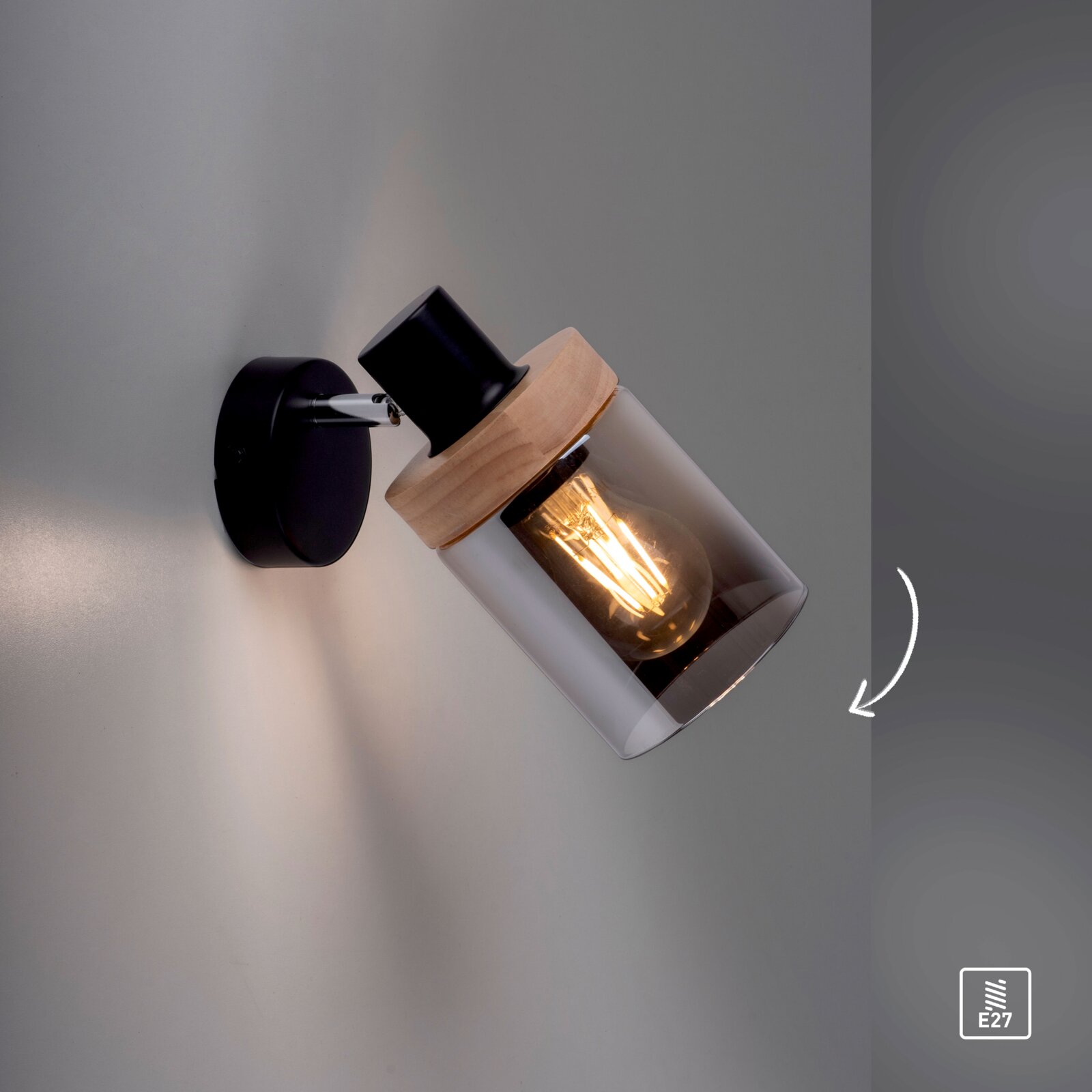 Moderne Wandlampe in Schwarz mit Holzdetail und zylindrischem Rauchglas-Schirm, leuchtendem Edison-Leuchtmittel (E27), schräg nach rechts ausgerichtet; Perspektive: seitliche Nahaufnahme in leichter Untersicht.