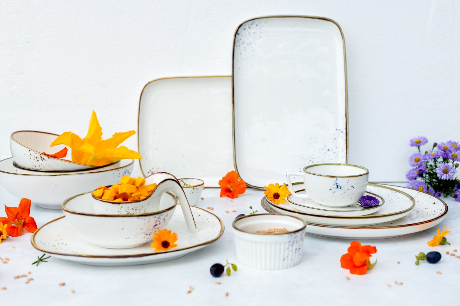Rustikales weißes Keramik-Geschirrset mit blauen Sprenkeln und braunem Rand (Teller, Schalen, Tasse) auf weißem Hintergrund mit Blumen und Früchten, Perspektive: leicht erhöhte Frontansicht