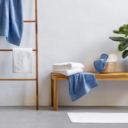 Handtuchset in Blau und Weiß, präsentiert auf einer Holzbank und einem Handtuchhalter, mit Pflanzen im Hintergrund, seitliche Perspektive.