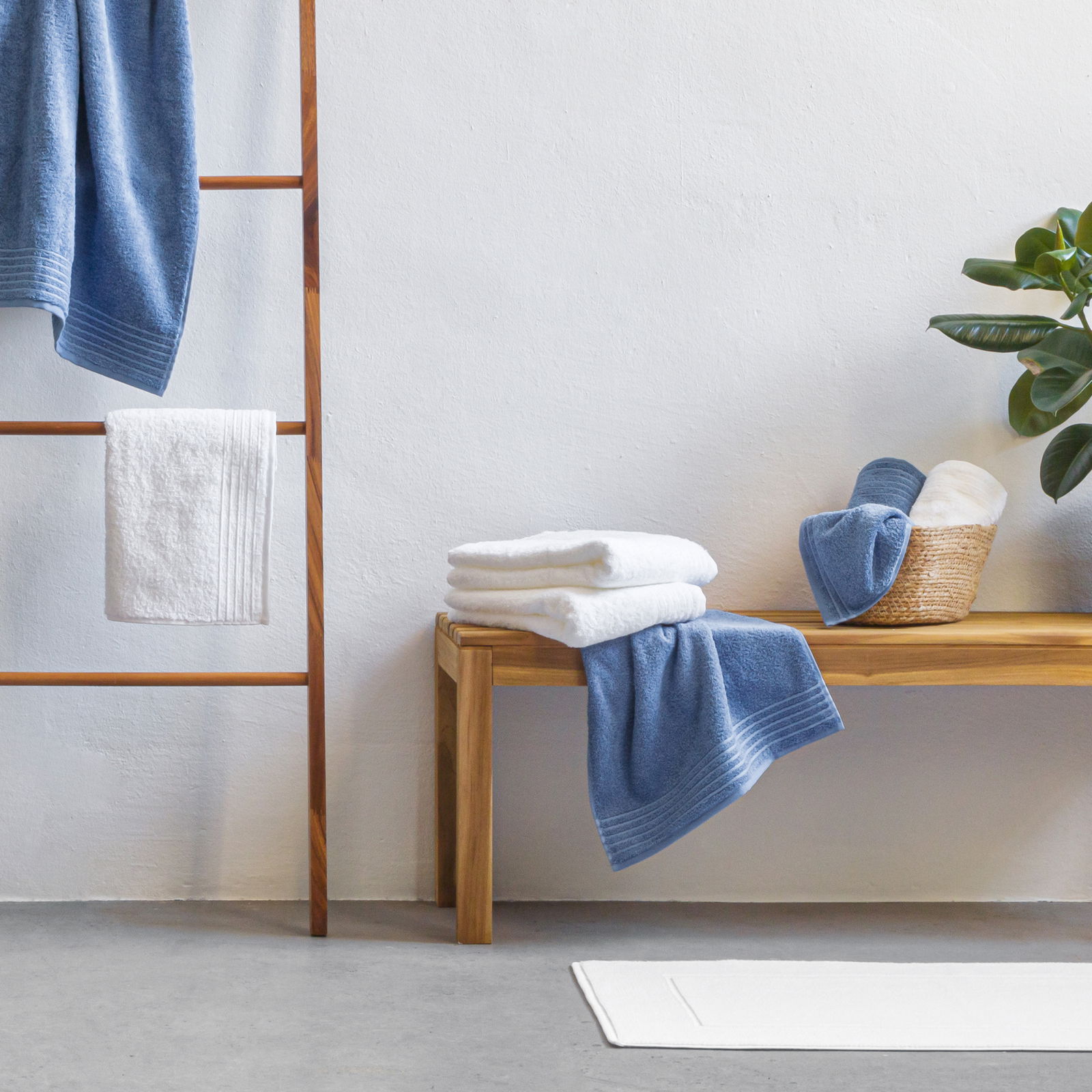 Handtuchset in Blau und Weiß, präsentiert auf einer Holzbank und einem Handtuchhalter, mit Pflanzen im Hintergrund, seitliche Perspektive.