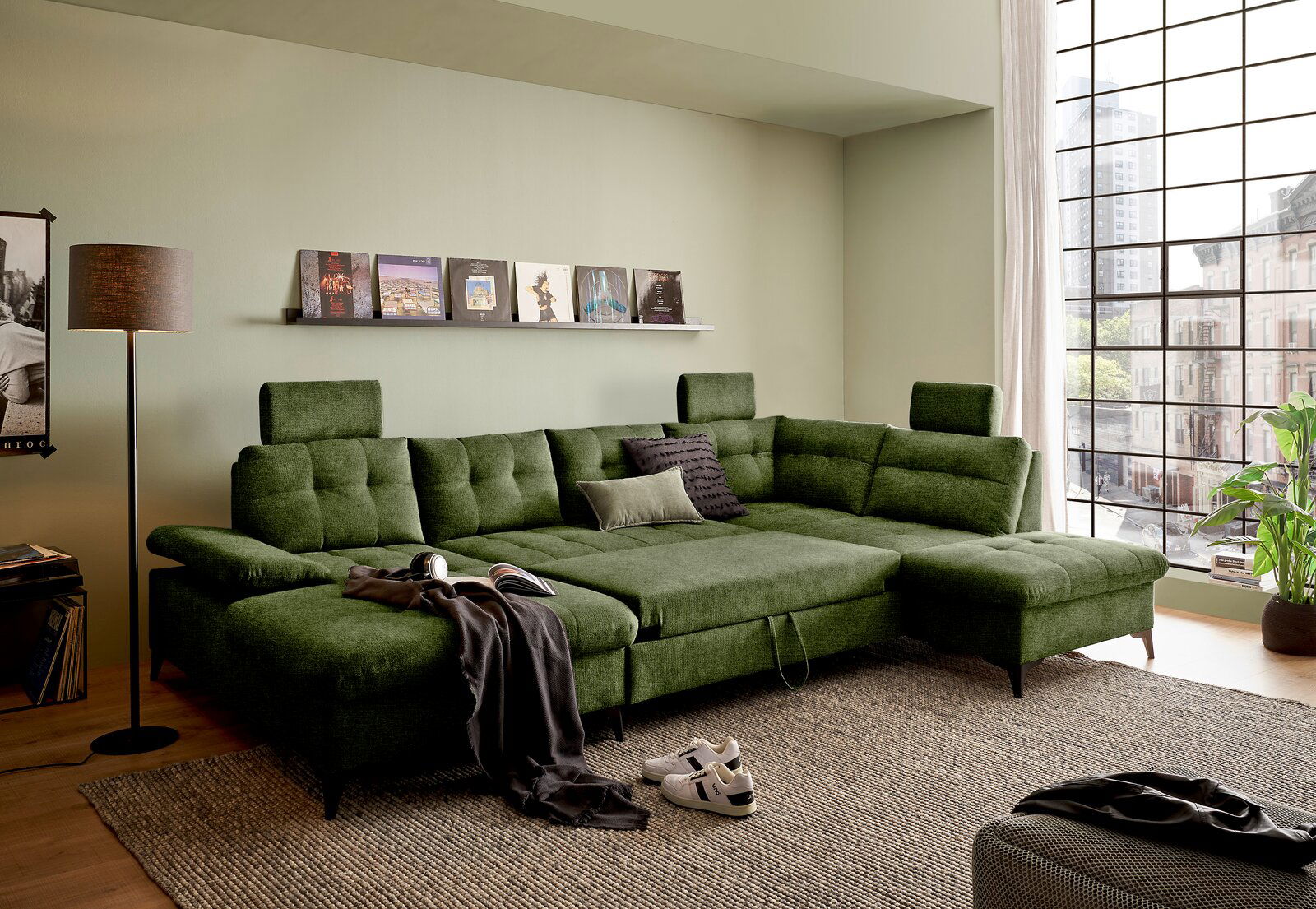 Grünes Ecksofa in einem modernen Wohnzimmer, fotografiert aus einer leicht erhöhten Perspektive. Das Sofa hat verstellbare Kopfstützen und ist mit Kissen dekoriert. Im Hintergrund sind ein Fenster mit Stadtblick und ein Regal mit Schallplatten zu sehen.