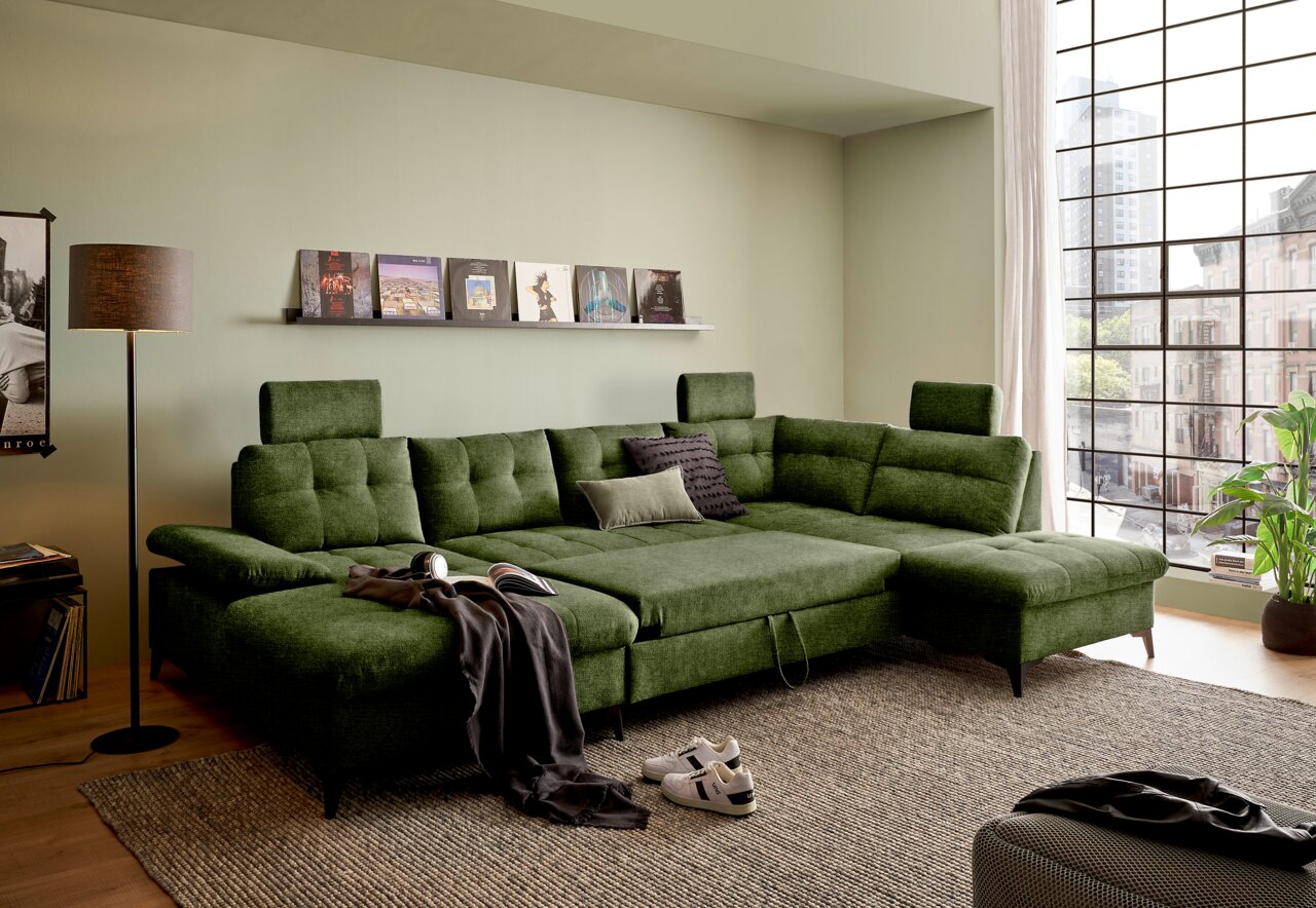 Grünes Ecksofa in einem modernen Wohnzimmer, fotografiert aus einer leicht erhöhten Perspektive. Das Sofa hat verstellbare Kopfstützen und ist mit Kissen dekoriert. Im Hintergrund sind ein Fenster mit Stadtblick und ein Regal mit Schallplatten zu sehen.