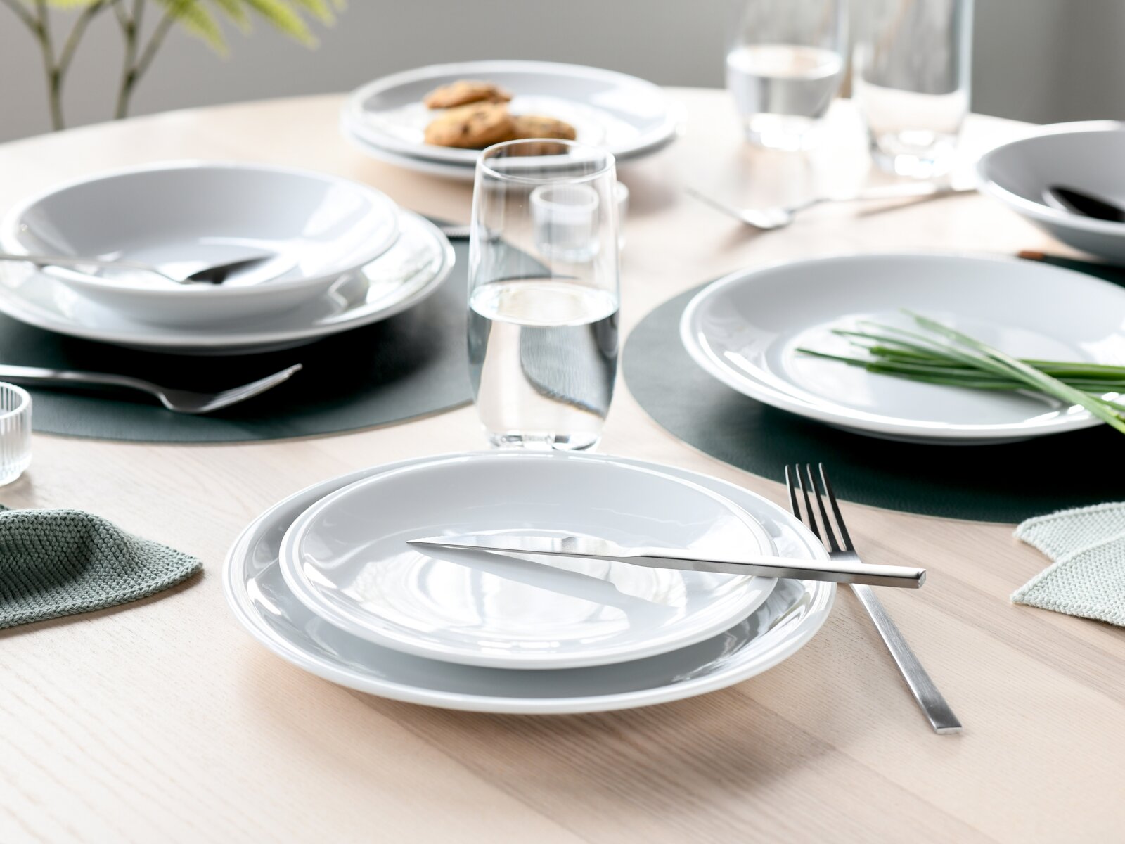 Modern gedeckter Esstisch mit weißen Tellern und Schalen, Edelstahlbesteck und Wassergläsern auf dunklen Platzsets, fotografiert aus leicht erhöhter, schräger Frontperspektive.