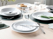 Modern gedeckter Esstisch mit weißen Tellern und Schalen, Edelstahlbesteck und Wassergläsern auf dunklen Platzsets, fotografiert aus leicht erhöhter, schräger Frontperspektive.