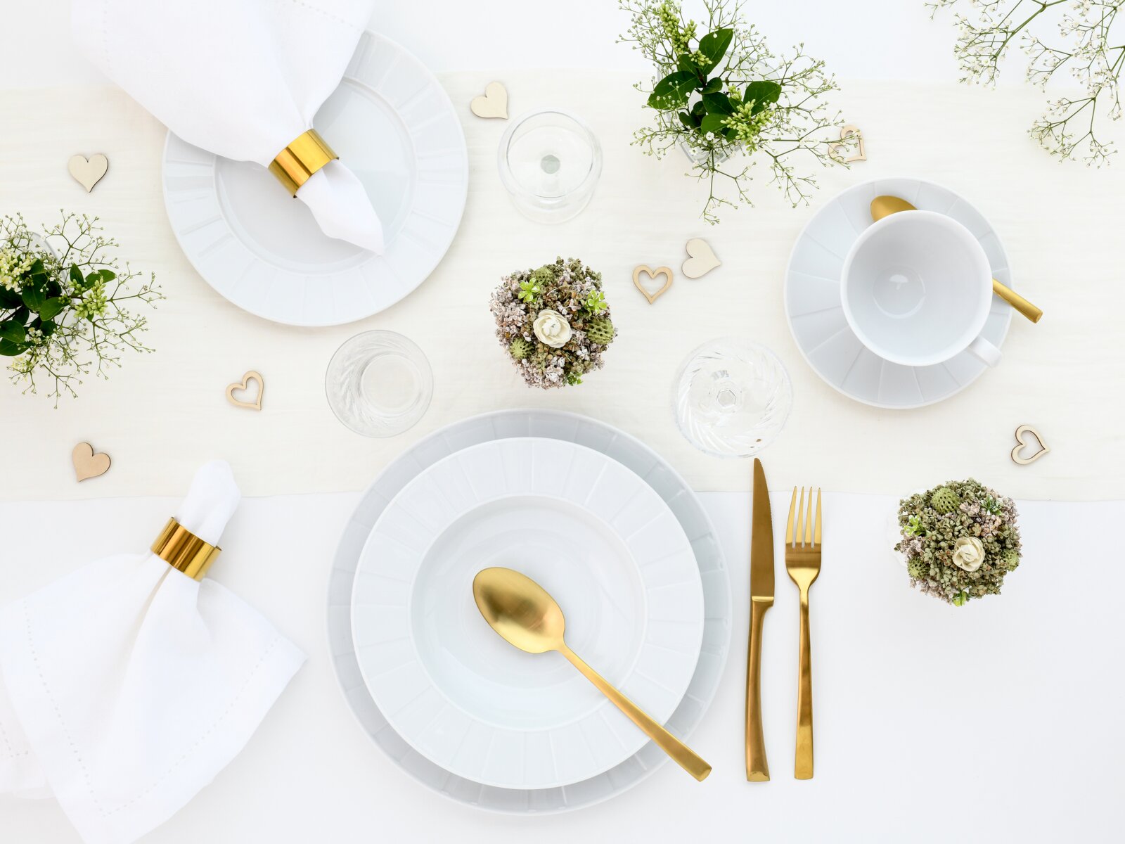 Festlich gedeckter Tisch in Draufsicht: weißes Geschirr mit goldfarbenem Besteck, Serviettenringen und kleinen Herzdekoren sowie grünen Blumenarrangements auf hellem Untergrund