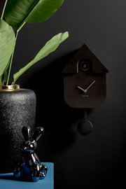 Moderne schwarze Wanduhr in Kuckucksuhrenform mit Pendel, seitliche Perspektive, dekoriert mit einer Pflanze und einer glänzenden Hasenfigur.