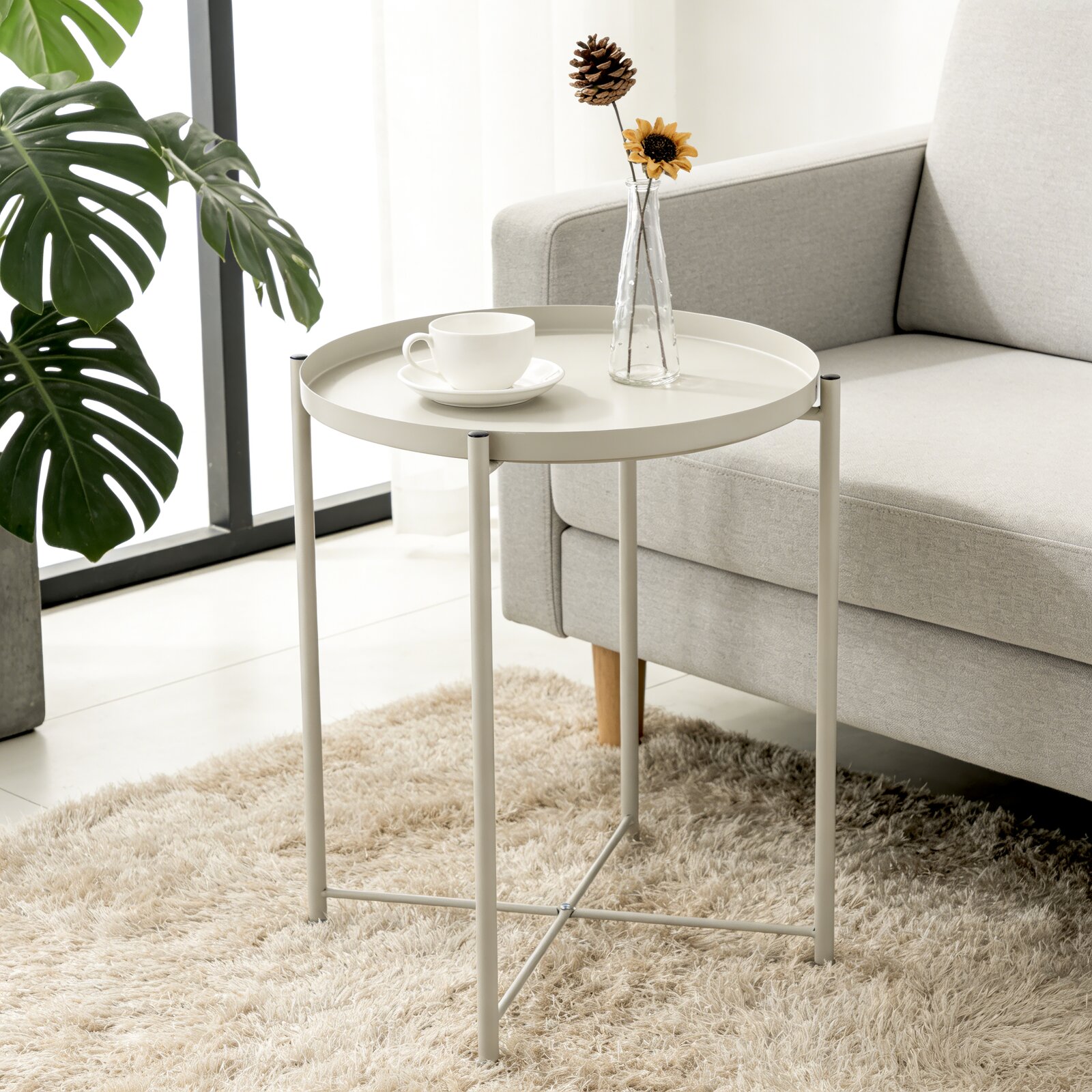 Carryhome Beistelltisch TIM Runder, hellgrauer Beistelltisch aus Metall neben einem grauen Sofa, mit Tasse und Glasvase mit Blume auf der Tischplatte, perspektivisch in leichter Draufsicht von vorne fotografiert.