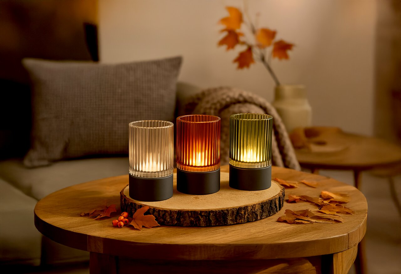 Drei Akku-Windlichter in verschiedenen Farben (klar, orange, grün) auf einem Holztisch, herbstlich dekoriert mit Blättern, aus einer frontalen Perspektive fotografiert.
