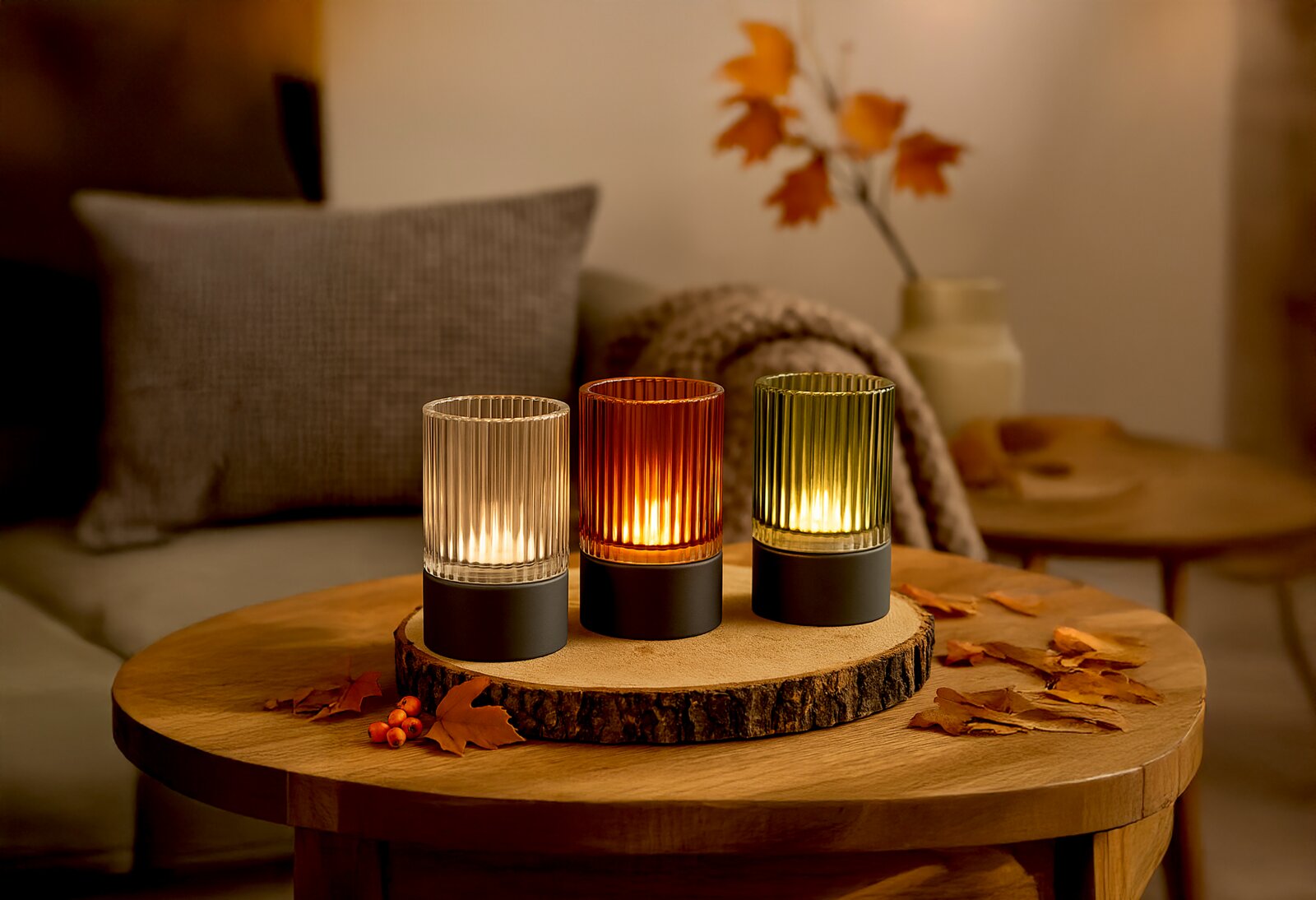 Drei Akku-Windlichter in verschiedenen Farben (klar, orange, grün) auf einem Holztisch, herbstlich dekoriert mit Blättern, aus einer frontalen Perspektive fotografiert.