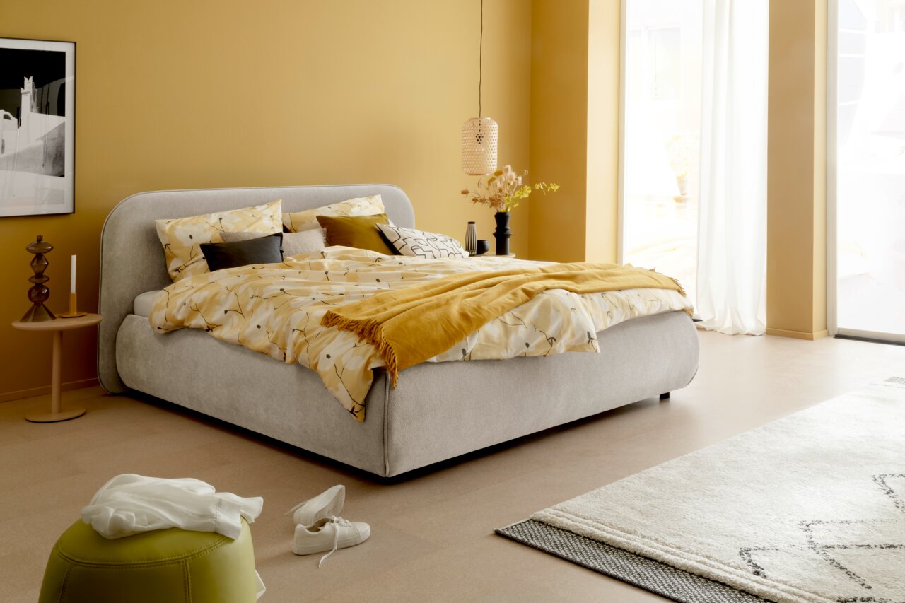 Modernes Polsterbettgestell in einem stilvollen Schlafzimmer, seitliche Perspektive. Das Bett ist mit gelber und gemusterter Bettwäsche dekoriert, umgeben von warmen, gelben Wänden und minimalistischen Möbeln.