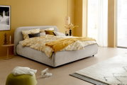 Een modern gestoffeerd bedframe in een stijlvolle slaapkamer, gezien vanaf de zijkant. Het bed is opgemaakt met geel beddengoed met een patroon en wordt omringd door warme, gele muren en minimalistische meubels.