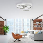 Moderne Deckenleuchte aus Stahl mit drei ineinander verschlungenen Ringen, von unten betrachtet in einem stilvollen Wohnzimmer mit großen Fenstern und Backsteinwänden.
