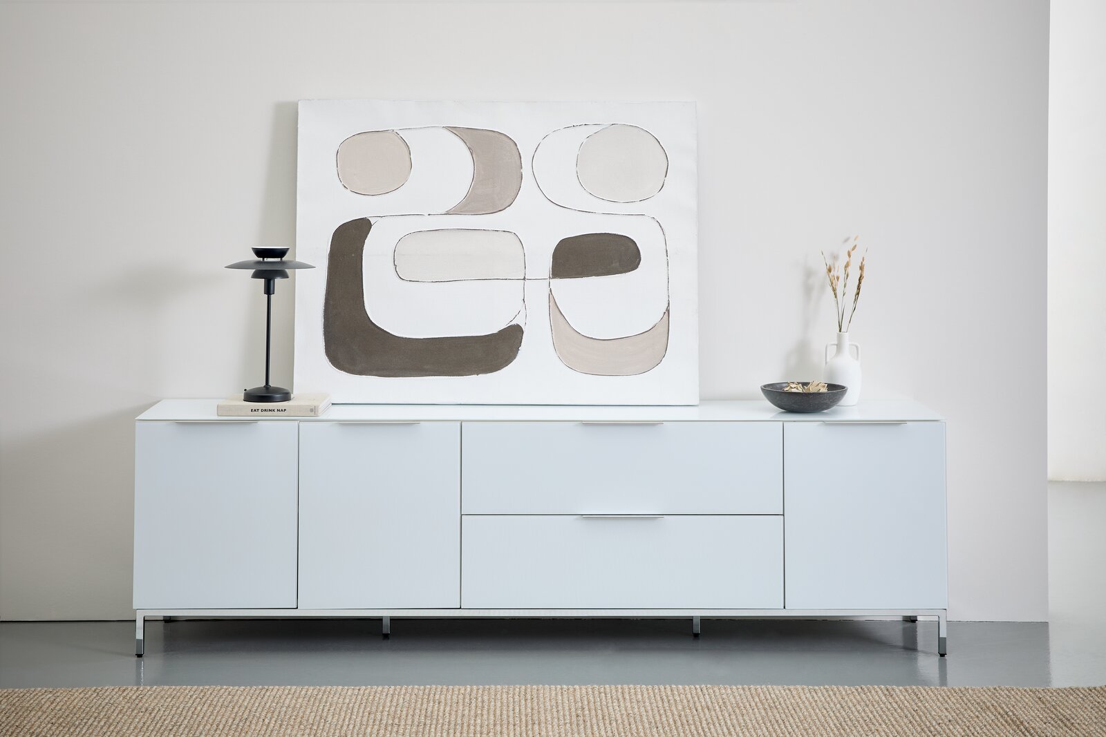 Frontale Ansicht eines modernen XL-TV-Lowboards in Weiß mit minimalistischer Dekoration, darunter eine abstrakte Kunstleinwand, eine schwarze Lampe und eine Vase.