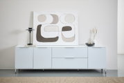 Frontale Ansicht eines modernen XL-TV-Lowboards in Weiß mit minimalistischer Dekoration, darunter eine abstrakte Kunstleinwand, eine schwarze Lampe und eine Vase.