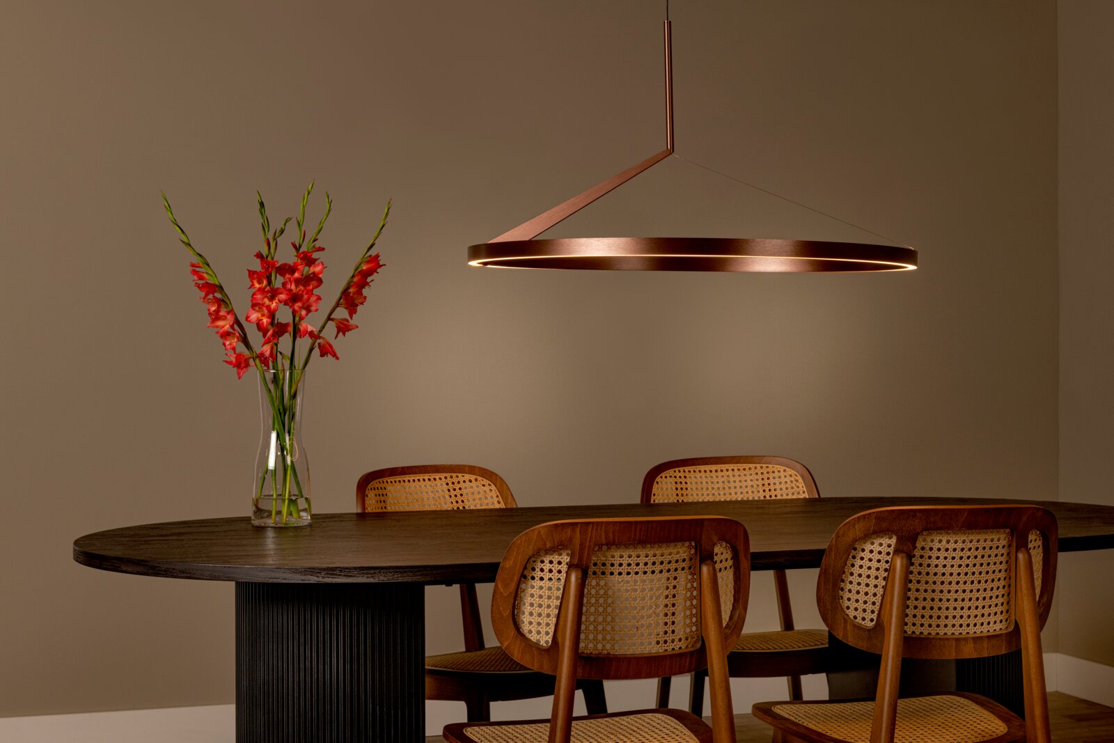 Moderne runde Pendelleuchte in Bronze/Kupferoptik mit warmem LED-Lichtring über einem dunklen Esstisch mit Holzstühlen und Blumenvase, Ansicht leicht seitlich von vorn