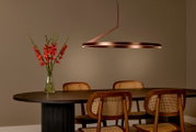 Moderne runde Pendelleuchte in Bronze/Kupferoptik mit warmem LED-Lichtring über einem dunklen Esstisch mit Holzstühlen und Blumenvase, Ansicht leicht seitlich von vorn
