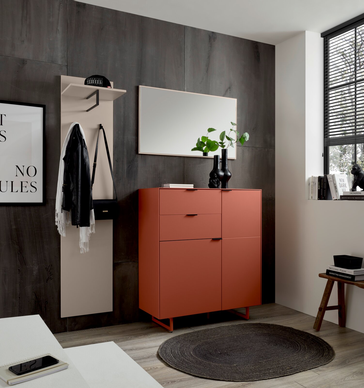 Voleo Schuhschrank ROSSO Modernes Sideboard in Terrakotta-Rot mit Schubladen und Türen auf Metallgestell, davor runder dunkler Teppich; Aufnahme aus leicht schräger Frontperspektive im Wohn-/Flurbereich mit Spiegel und Garderobe an dunkler Wand.