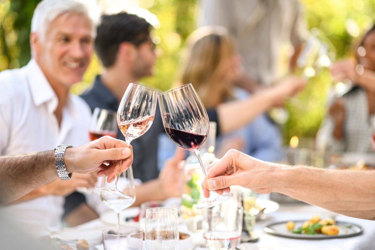Zwei Personen stoßen mit Bordeauxgläsern bei einem geselligen Treffen im Freien an, im Hintergrund sind weitere Menschen zu sehen.