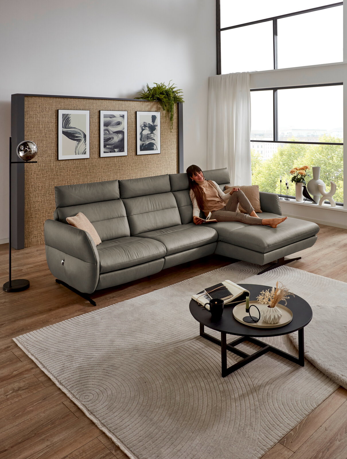Dieter Knoll Longchairkombination MATINO Graue Longchairkombination MATINO von Dieter Knoll in einem modernen Wohnzimmer mit großen Fenstern, einer Frau, die entspannt auf dem Sofa liegt, und einem runden Couchtisch im Vordergrund. Perspektive von vorne.