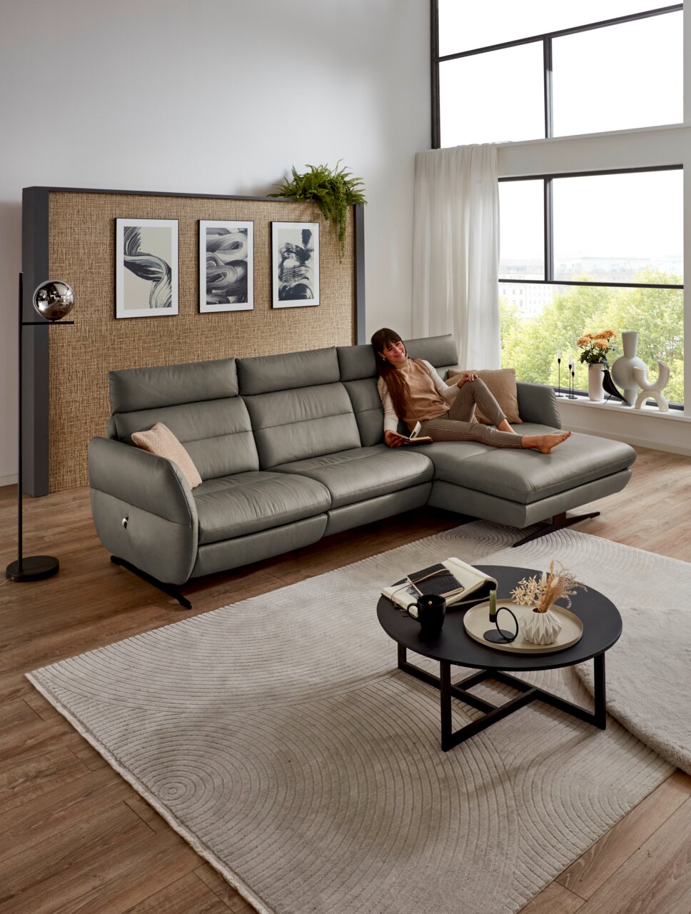 Graue Longchairkombination MATINO von Dieter Knoll in einem modernen Wohnzimmer mit großen Fenstern, einer Frau, die entspannt auf dem Sofa liegt, und einem runden Couchtisch im Vordergrund. Perspektive von vorne.