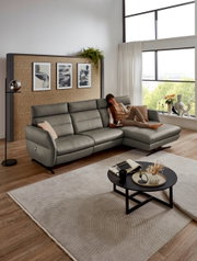 Grijze MATINO-longchaircombinatie van Dieter Knoll in een moderne woonkamer met grote ramen, een vrouw die ontspannen op de bank ligt en een ronde salontafel op de voorgrond. Aanzicht van voren.