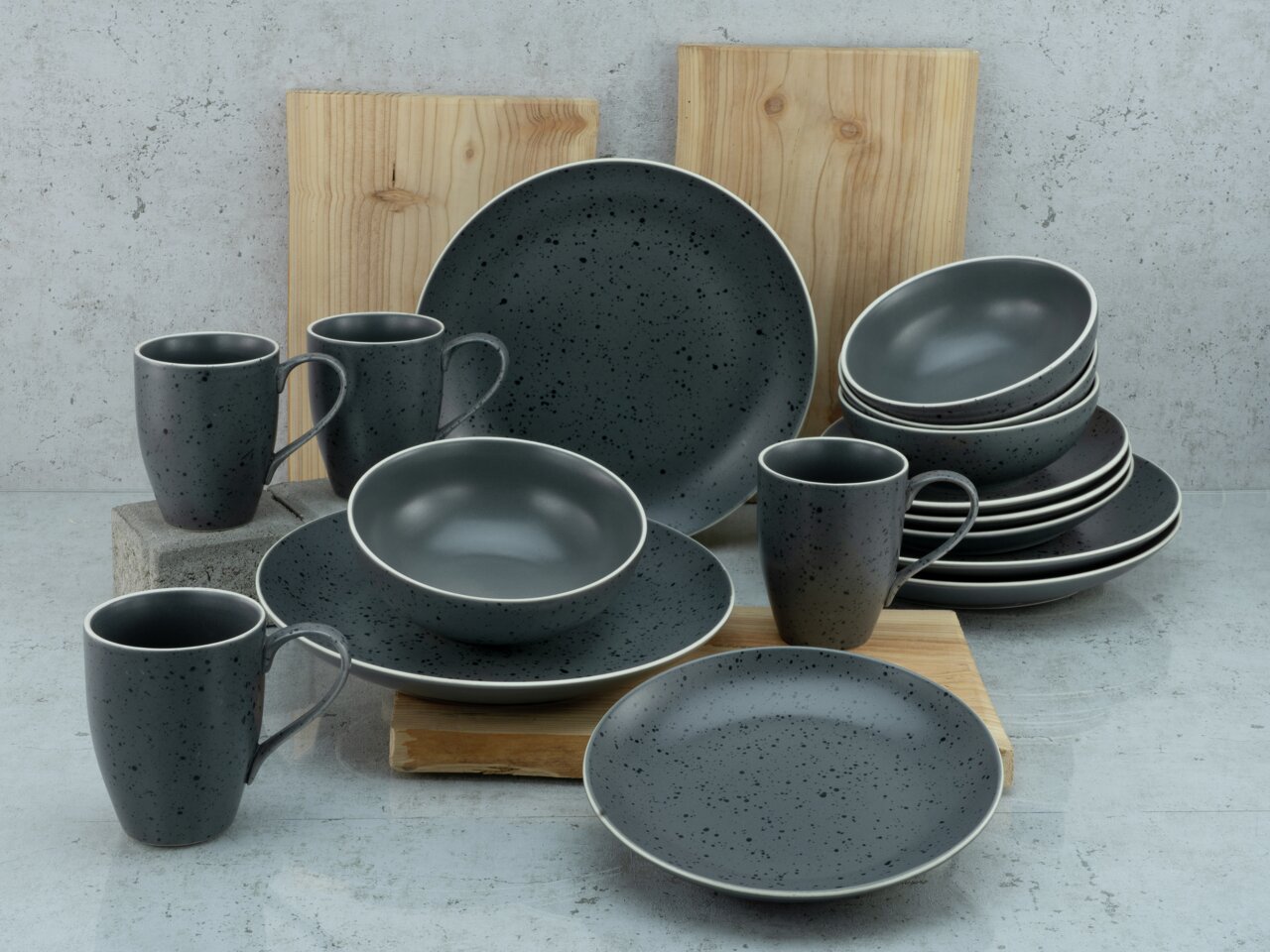 creaTable Kombiservice 16-tlg. INDUSTRIAL CHIC Kombiservice Industrial Chic in dunklem, gesprenkeltem Design, bestehend aus Tassen, Tellern und Schalen, auf einer Betonoberfläche mit Holzplatten im Hintergrund, seitliche Perspektive.