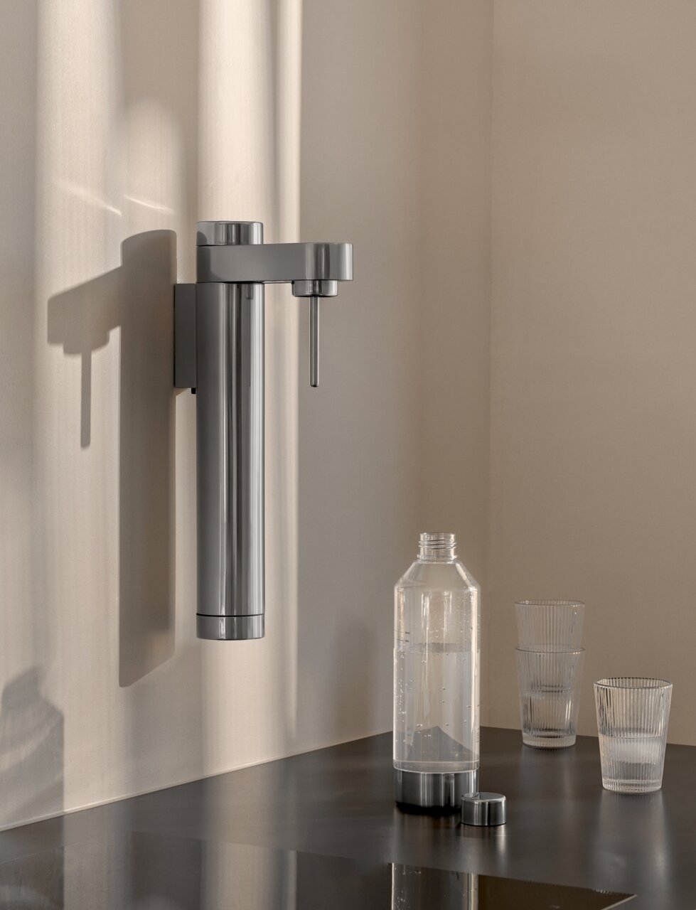 Stelton Wassersprudler BRUS Moderner Wand-Wassersprudler aus Edelstahl mit Flasche und Gläsern auf einer Arbeitsplatte, seitliche Perspektive.