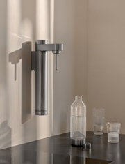 Moderner Wand-Wassersprudler aus Edelstahl mit Flasche und Gläsern auf einer Arbeitsplatte, seitliche Perspektive.
