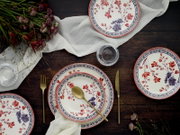 Draufsicht auf einen gedeckten Holztisch mit floral gemustertem Keramikgeschirr in Weiß mit roten und blauen Blumen, goldfarbenem Besteck, Leinenserviette, Gläsern und Blumenarrangement.