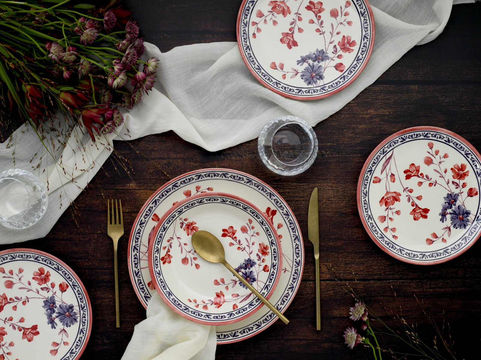 Draufsicht auf einen gedeckten Holztisch mit floral gemustertem Keramikgeschirr in Weiß mit roten und blauen Blumen, goldfarbenem Besteck, Leinenserviette, Gläsern und Blumenarrangement.