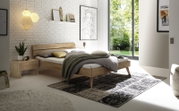 Modernes Schlafzimmer mit einem Bett aus Holz und Metall, seitliche Perspektive. Das Bett ist mit grauer Bettwäsche und Kissen dekoriert, daneben ein Nachttisch mit Pflanze und Lampe. Große Fenster lassen viel Tageslicht herein.