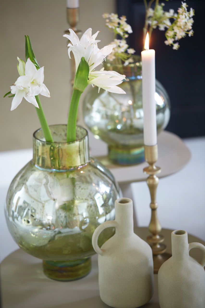 Große, runde Glasvase mit weißen Blumen, umgeben von Kerzen und Keramikvasen, aus einer leicht erhöhten Perspektive fotografiert.