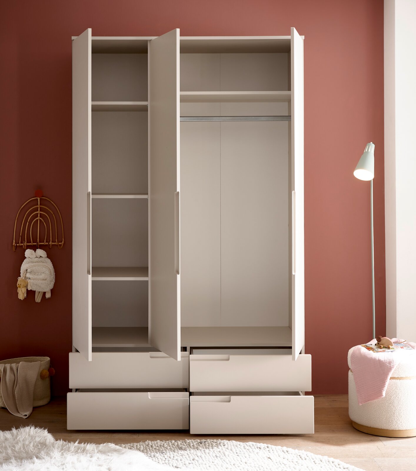 Weißer Kleiderschrank mit drei geöffneten Türen, Innenfächern und Kleiderstange sowie vier Schubladen unten, fotografiert in Frontalansicht leicht von links in einem Kinderzimmer.