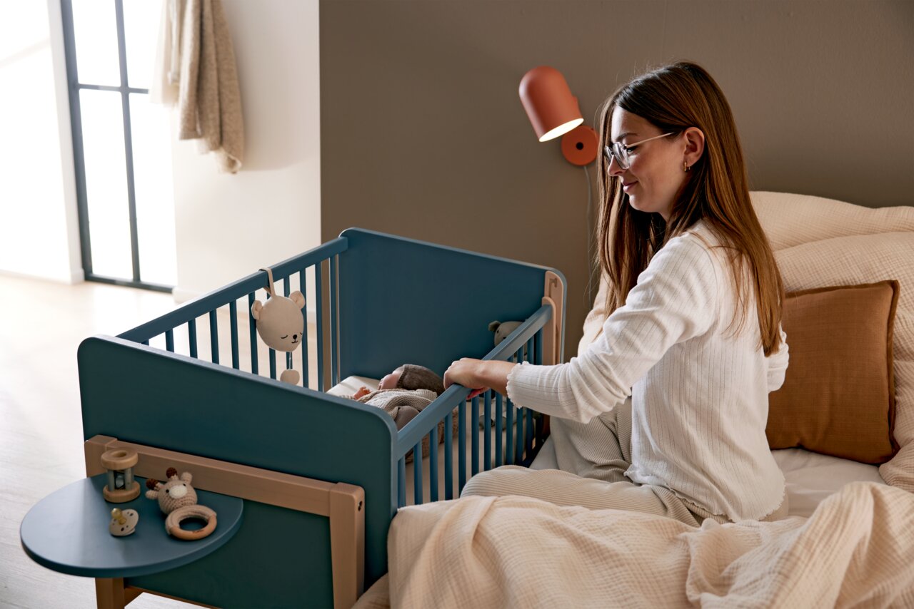 Frau sitzt auf einem Bett und schaut in ein blaues Babybett mit Spielzeug, seitliche Perspektive.