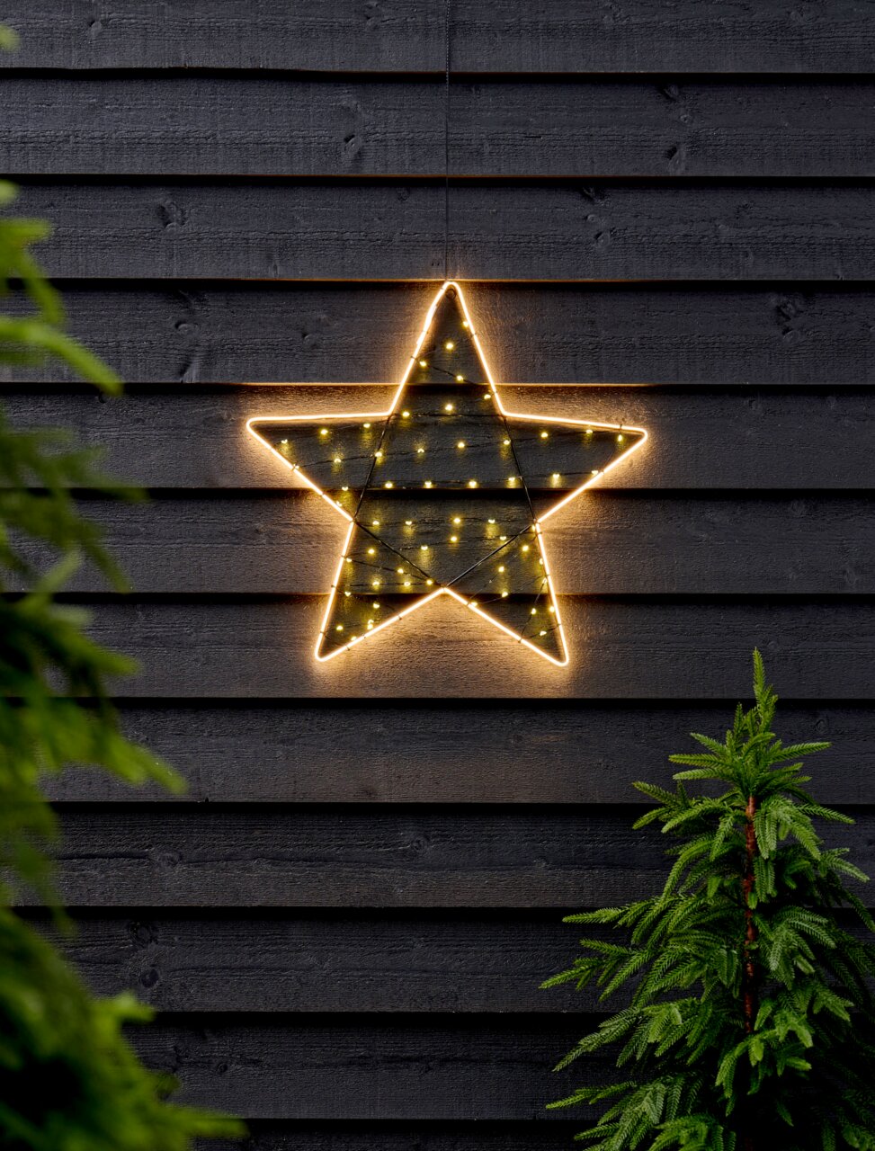 Leuchtender Stern mit Timer, 47 cm, an einer dunklen Holzwand hängend, frontal fotografiert, umgeben von grünen Tannen.