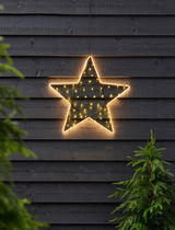 Leuchtender Stern mit Timer, 47 cm, an einer dunklen Holzwand hängend, frontal fotografiert, umgeben von grünen Tannen.