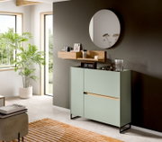 Modernes Sideboard in Salbeigrün mit schwarzem Metallgestell und Holzdetails, in einem hellen Wohnraum unter einem runden Wandspiegel – Perspektive: leicht schräg von vorn rechts.