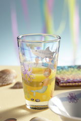 Kinderglas mit Meeresmotiven, darunter ein Hai, eine Krake und ein Kind als Meerjungfrau, gefüllt mit gelbem Saft, aus seitlicher Perspektive fotografiert.